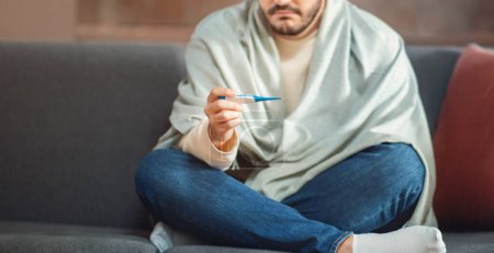 Cropped of man appears to be feeling unwell, sitting on a gray couch with a blanket wrapped around his shoulders while holding a thermometer, possibly checking for a fever