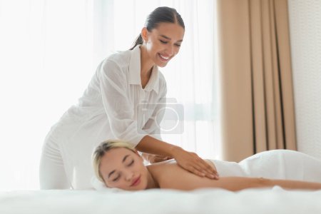 A woman is lying face down on a massage table while a massage therapist is applying pressure to her back in a spa setting, copy space