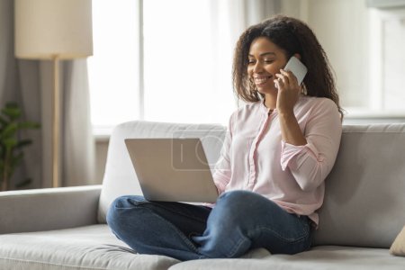 Téléchargez les photos : Cette image représente une jeune femme noire sur un canapé, profondément engagée dans une conversation téléphonique tout en travaillant simultanément sur son ordinateur portable, mettant en valeur un environnement dynamique de travail à domicile - en image libre de droit