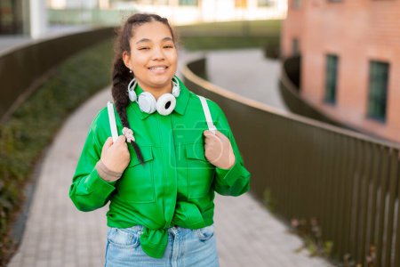 Foto de Una joven estudiante con una chaqueta verde y auriculares está de pie al aire libre. Ella aparece enfocada mientras escucha música, posiblemente disfrutando de un paseo o participando en actividades al aire libre. - Imagen libre de derechos