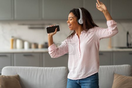Glückliche junge schwarze Frau mit Kopfhörern, die zu Hause singt und tanzt und das Smartphone als Mikrofon benutzt