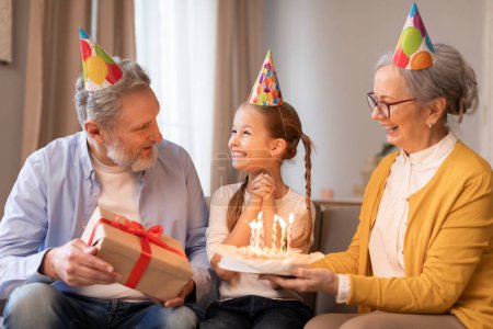 Foto de A young girl sits between her smiling grandparents who are both wearing party hats. The grandfather holds a wrapped gift with a red bow, while the grandmother presents her with a birthday cake - Imagen libre de derechos