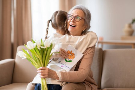 Photo for A cheerful elderly woman is sitting on a couch, warmly hugging a little girl who is presenting her with a bouquet of white tulips and a handmade card - Royalty Free Image