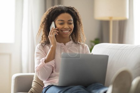 Foto de Capturada en una acogedora sala de estar, esta foto muestra a una mujer afroamericana enfocada hablando por teléfono mientras trabaja en su computadora portátil, encarnando un entorno productivo de oficina en casa. - Imagen libre de derechos