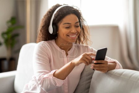 This image portrays a content African American woman relaxing on her sofa, enjoying music through her headphones and phone, creating a tranquil atmosphere in a modern home environment