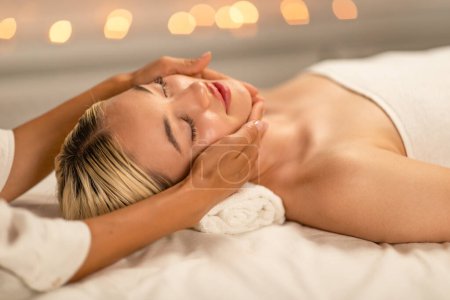 A serene lady lies on her back, eyes closed, with a peaceful expression as she receives a facial massage. Soft lighting and blurred lights in the background create an atmosphere of tranquility
