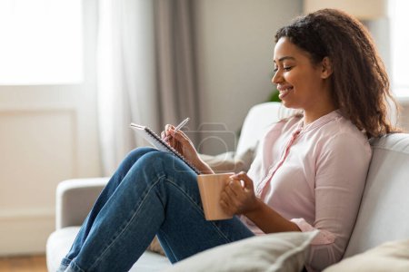 Radiant de positivité, une jeune afro-américaine profite de son café matinal tout en écrivant des pensées dans son journal, confortablement installé sur son canapé