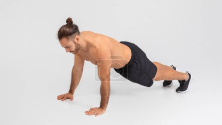 A man is doing push ups on a plain white background. He appears to be in the middle of a workout routine, using his arms to lower and raise his body in a repetitive motion.