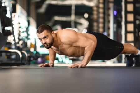 Un hombre con atuendo atlético está haciendo flexiones en el piso del gimnasio. Los músculos del hombre están visiblemente comprometidos a medida que baja y levanta su cuerpo con movimientos controlados