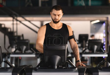 A man wearing athletic clothing is jogging on a treadmill machine inside a bright and modern gym. The equipment is surrounded by other exercise machines and gym-goers in the background.
