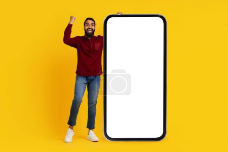 Téléchargez les photos : Un Indien barbu avec un large sourire lève le poing dans un geste de célébration à côté d'un modèle géant de smartphone avec un écran blanc, sur fond jaune vif - en image libre de droit