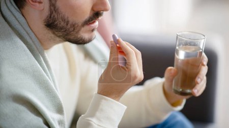 Gestutzt von einem bärtigen Mann, der eine Pille zwischen seinen Fingern hält, bereit, sie mit Hilfe eines vollen Glases Wasser zu schlucken, was einen Moment persönlicher Gesundheitsfürsorge hervorhebt