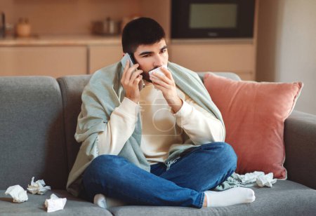A young man, wrapped in a blanket, sits on his couch surrounded by tissues, indicating he is unwell. He holds a tissue in one hand while talking on the phone, possibly seeking medical advice