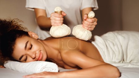Afroamerikanerin liegt mit dem Gesicht nach unten und erhält eine traditionelle Kräuterkompresse-Massage von einer Masseurin in einer ruhigen Wellness-Umgebung