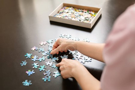 Les mains des femmes organisent méticuleusement de petites pièces de puzzle bleu et blanc sur une surface noire élégante, avec une boîte de pièces supplémentaires à proximité, montrant un moment de concentration et de résolution de problèmes