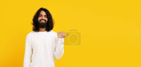 Indian man with long hair and a beard is depicted wearing a white shirt, pointing at copy space isolated on yellow background, panorama