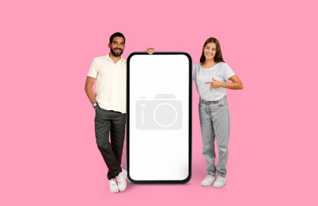 Photo for A young man and woman leaning and pointing at an oversized smartphone screen on a pink backdrop, mockup - Royalty Free Image