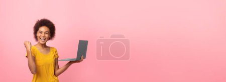 A cheerful young african american woman with curly hair is standing against a pink background, holding a laptop and celebrating or showing success