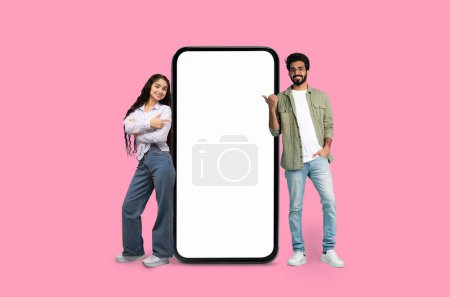 Ein junger indischer Mann und eine junge Frau präsentieren fröhlich eine Smartphone-Attrappe auf rosa Studiohintergrund
