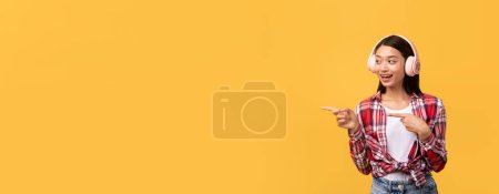 Photo for A joyful young asian woman wearing a plaid shirt and headphones is pointing to her right with a cheerful expression against a yellow background - Royalty Free Image