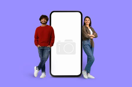 Joven pareja india apoyándose casualmente en un enorme teléfono inteligente con una pantalla blanca sobre un telón de fondo púrpura