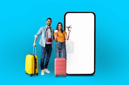 Photo for Young multiethnic couple with suitcases stands next to an empty smartphone screen, highlighting application use for travel, isolated on a blue background - Royalty Free Image