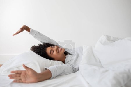 Hispanic woman is lying in bed with her arms stretched out to the sides. She appears relaxed and comfortable, possibly waking up or winding down for the day.
