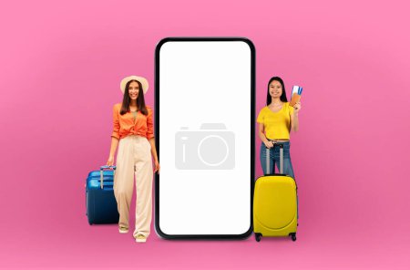 Téléchargez les photos : Deux femmes avec des valises se tiennent à côté d'un grand smartphone, indiquant une application de voyage et de vacances ou une offre en ligne liée à la technologie numérique. Ils sont isolés sur un fond rose - en image libre de droit