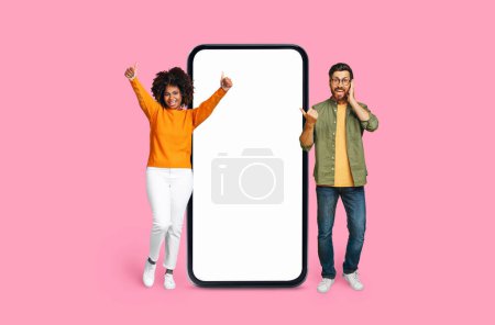 Joyful Multiracial man and woman with energetic poses next to a large smartphone screen on a pink backdrop