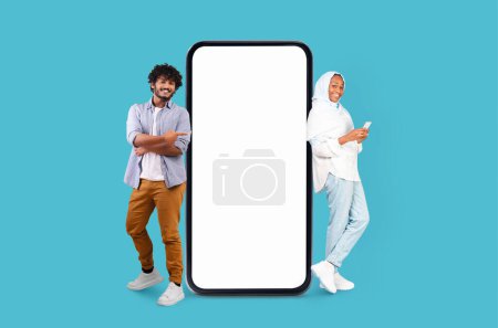 Téléchargez les photos : Homme et femme multiraciaux en vêtements décontractés posant aux côtés d'une maquette géante de smartphone sur un fond bleu - en image libre de droit
