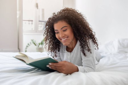 Femme hispanique couchée confortablement sur un lit, absorbée par un livre qu'elle lit. La scène capture sa posture détendue et focalise l'attention sur les pages.