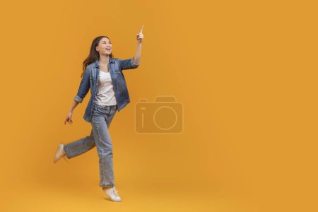 Excited young asian woman running and pointing upwards, isolated on an orange background, copy space