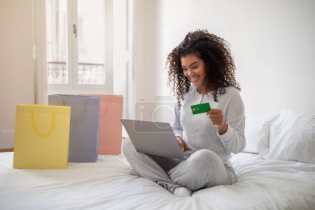 Téléchargez les photos : AHispanicwoman est assise sur un lit, tenant une carte de crédit et utilisant un ordinateur portable. Elle semble se concentrer sur l'écran, peut-être faire un achat en ligne ou gérer les finances. - en image libre de droit