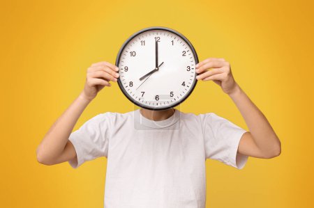 Photo for Time management concept. Teen boy covering his face with big clock, orange panorama background - Royalty Free Image