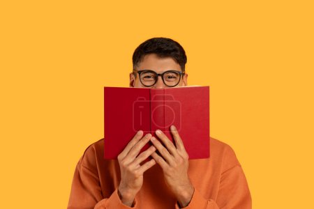 A man is seen holding a red book over his face, covering his features. The book stands out against his skin, creating a contrast in colors.