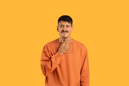 A young man sports a casual mustache and a thoughtful expression while wearing a bright orange sweater. He stands with one hand gently touching his neck, suffering from throat ache