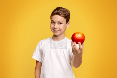 Foto de Niño positivo que ofrece manzana roja fresca en la cámara, fondo de estudio naranja - Imagen libre de derechos