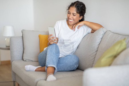 Foto de Una mujer está sentada en un sofá, sonriendo mientras mira atentamente la pantalla de su teléfono celular. Ella aparece relajada y comprometida con el contenido de su dispositivo. - Imagen libre de derechos