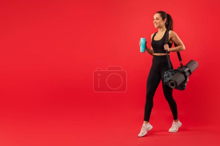 Téléchargez les photos : Une femme portant un haut de soutien-gorge de sport noir et leggings est représentée tenant une tasse bleue dans sa main, marchant vers l'espace de copie, isolé sur fond rouge - en image libre de droit