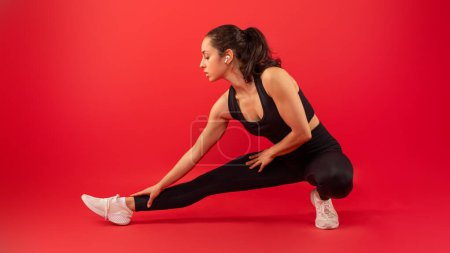 A woman wearing a black top and leggings is shown stretching her arms and legs in a fitness routine. The black clothing contrasts with her skin, emphasizing her movements as she warms up