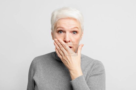 Eine schockierte ältere europäische Frau, die ihren Mund vor grauem Hintergrund verhüllt und das Erstaunen über s3niorlife beschreibt