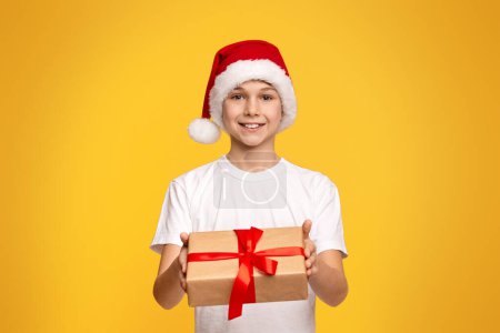 Foto de Felices fiestas. Lindo adolescente en Santa sombrero ofreciendo caja de regalo en la cámara, fondo de estudio naranja - Imagen libre de derechos