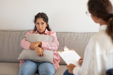 Una joven parece pensativa mientras sostiene un cojín cerca mientras está sentada en un sofá gris, frente a una terapeuta que sostiene un portapapeles y toma notas durante una sesión de terapia.