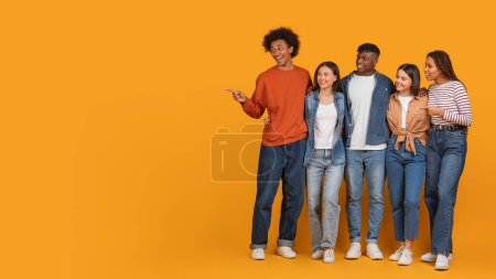 Foto de Cinco diversos adultos jóvenes sonríen y miran a la derecha mientras un hombre apunta en esa dirección sobre un fondo amarillo, copiando espacio - Imagen libre de derechos