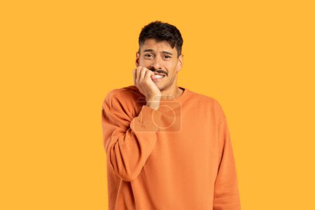 Photo for Confused man wearing an orange sweater is standing with his hand held up to his face, appearing contemplative or upset. He has a pensive expression, deep in thought. - Royalty Free Image