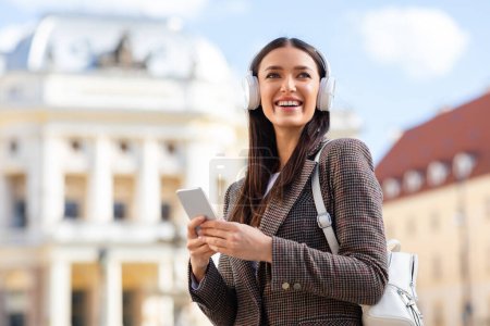 A millennial lady smiling while using a smartphone and headphones outdoors, set against a European city scene