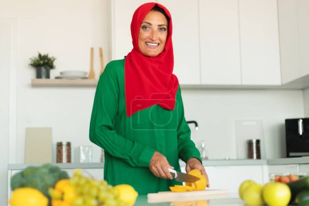 Foto de Una mujer que lleva un hiyab rojo y verde está cortando hábilmente un pimiento en una tabla de cortar de madera. Ella está enfocada en su tarea, rebanando delicadamente el queso con precisión. - Imagen libre de derechos