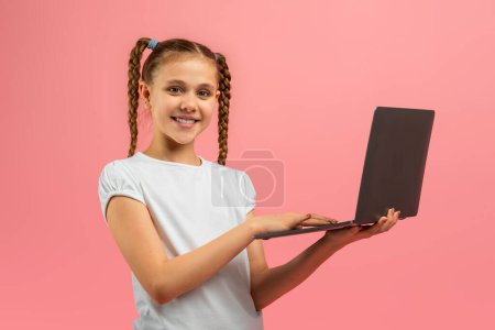 Ein fröhliches junges Mädchen mit geflochtenen Haaren steht vor rosa Hintergrund und hält stolz einen offenen Laptop