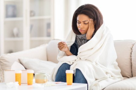 Photo for African American woman is seated on a couch, with a cozy blanket draped over her head, holding thermometer, suffering from fever - Royalty Free Image