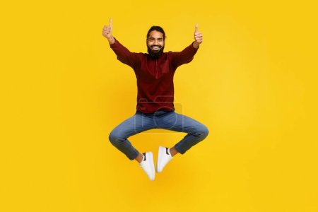 A beaming Indian man with a beard is captured mid-jump, giving two enthusiastic thumbs up. He is casually dressed in a burgundy sweater and jeans